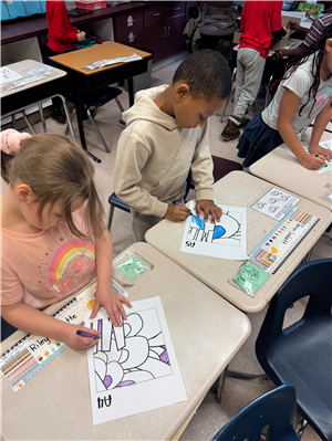 Photo shows two female students working at a desk, coloring in pages that will be put together to form a bigger picture.